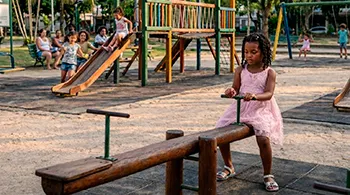 Menina brincando sozinha em um parquinho infantil enquanto outras crianças brincam juntas ao fundo, ilustrando reflexões sobre pertencimento e questões raciais na infância.